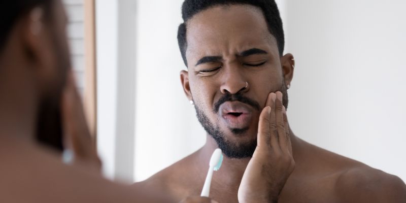 man who's holding his jaw in pain with one hand and a tooth brush in another